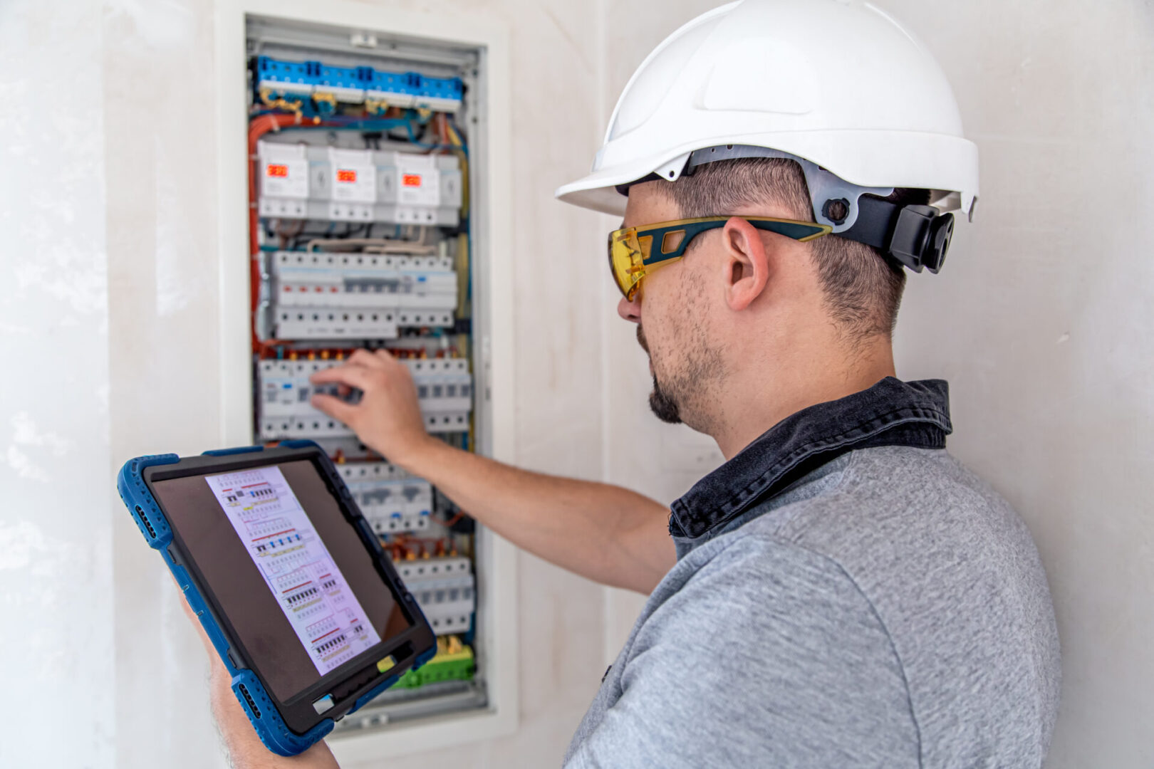 A worker in a hard hat checks an electrical panel, meticulously examining the connections while holding a tablet displaying data. Nearby, tools for outlet installation lie ready for the next task.