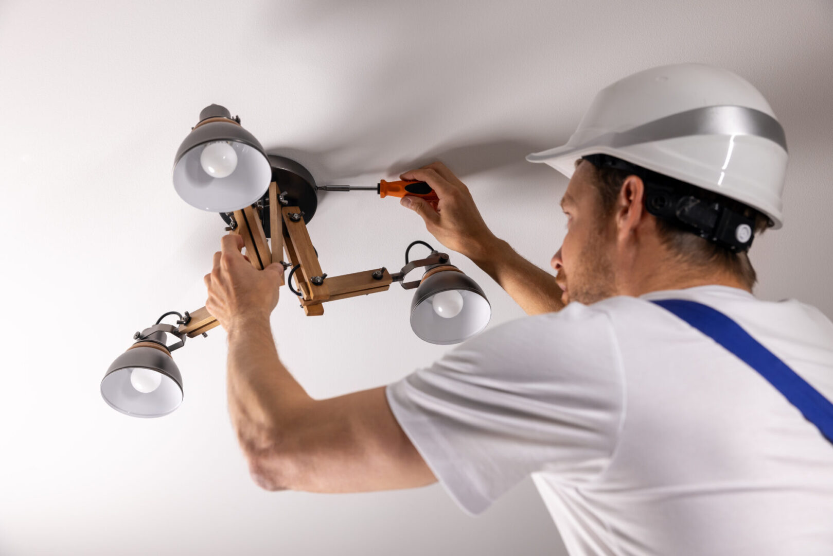 A person in a hard hat and overalls expertly uses a screwdriver to carry out custom electrical work, installing or repairing a ceiling light fixture with multiple bulbs.