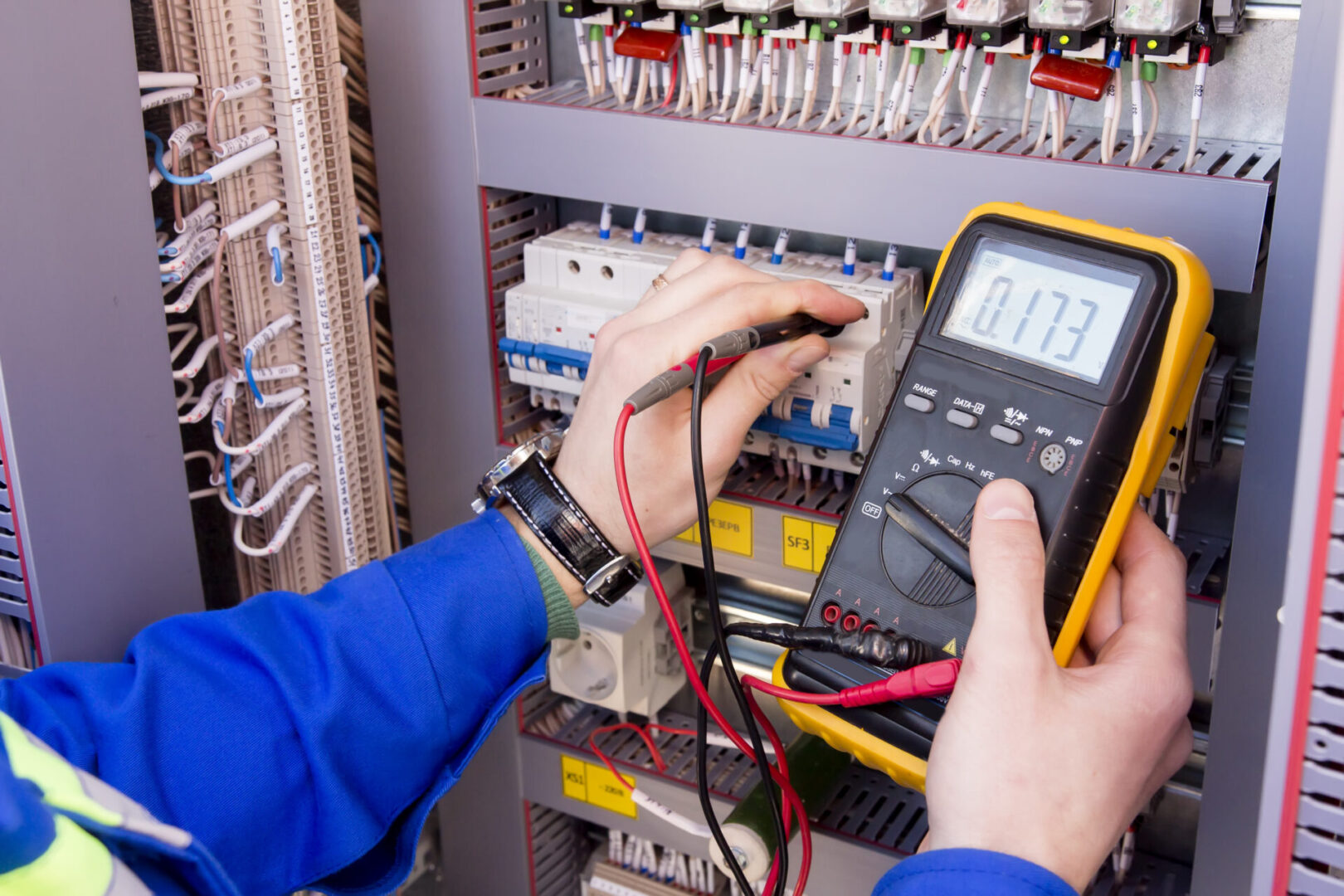 A person expertly uses a multimeter to measure voltage in an electrical panel, showcasing their skills in custom electrical work, with wires and circuit breakers clearly visible.